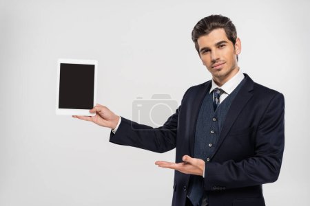 jeune homme d'affaires en costume pointant vers tablette numérique avec écran blanc isolé sur gris 