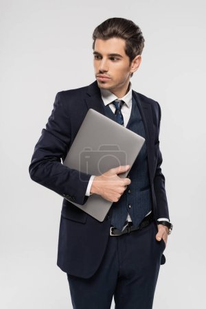 joven hombre de negocios en traje que sostiene el ordenador portátil y de pie con la mano en el bolsillo aislado en gris