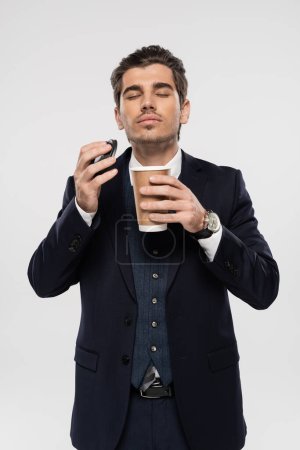 hombre de negocios con los ojos cerrados sosteniendo taza de papel y tapa mientras huele café de la mañana aislado en gris