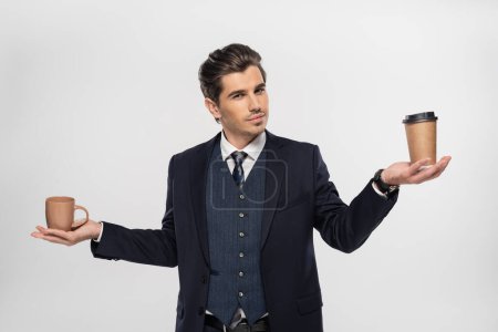young businessman holding paper cup and mug while making choice isolated on grey