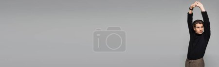 Téléchargez les photos : Jeune homme beau en col roulé noir posant avec les mains levées isolé sur gris, bannière - en image libre de droit