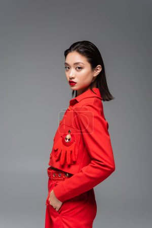 modelo asiático morena en chaqueta roja decorada con guante posando con mano en bolsillo aislado en gris