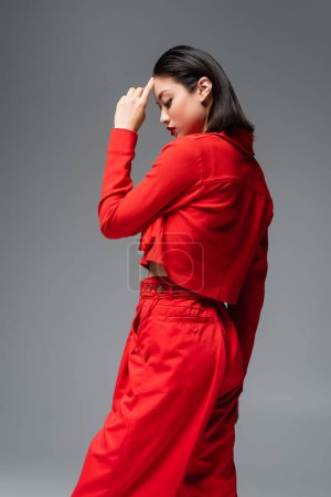 Photo for Side view of asian woman in red stylish attire touching forehead isolated on grey - Royalty Free Image