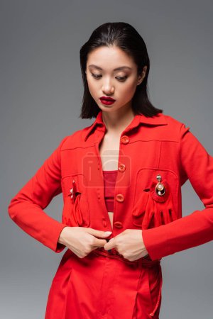joven mujer asiática en chaqueta roja decorada con broches y guantes aislados en gris