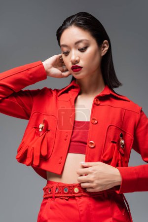 modelo asiático joven en blazer rojo decorado con broches y guantes aislados en gris