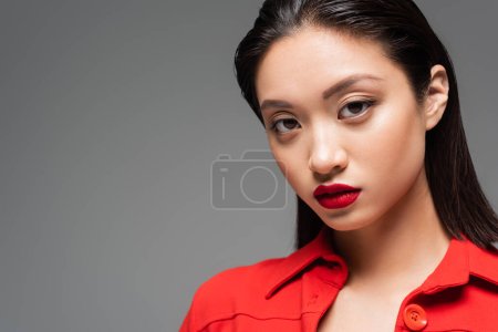 portrait of brunette asian woman with makeup and red lips looking at camera isolated on grey