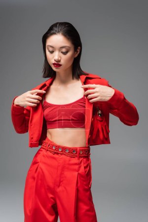 Photo for Brunette asian woman in top and pants taking off red jacket isolated on grey - Royalty Free Image