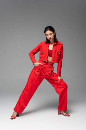 volle Länge der asiatischen Frau im roten eleganten Anzug posiert mit der Hand auf der Hüfte auf grauem Hintergrund