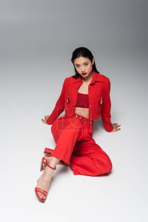 sensual asian woman in red suit and heeled sandals sitting on grey background