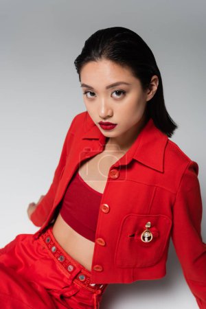 trendige asiatische Frau in roter Jacke mit Brosche sitzt und blickt in die Kamera auf grauem Hintergrund