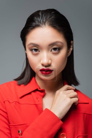 portrait of brunette asian woman with red lips wearing red blazer and looking at camera isolated on grey