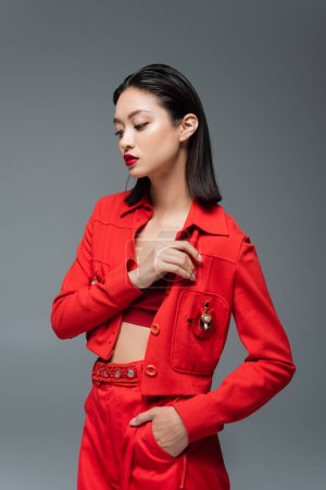asian woman in red pants and jacket with brooch posing with hand in pocket isolated on grey