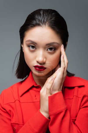 portrait of pretty asian woman in red fashionable jacket holding hands near face isolated on grey