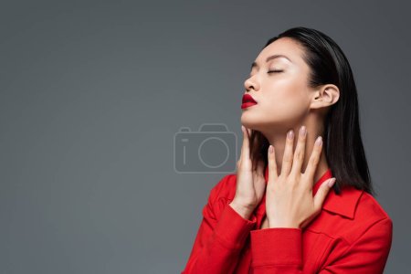 sensual asian woman with closed eyes touching neck while posing in red jacket isolated on grey