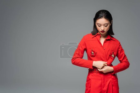 Foto de Morena mujer asiática tocando chaqueta roja decorada con broches aislados en gris - Imagen libre de derechos