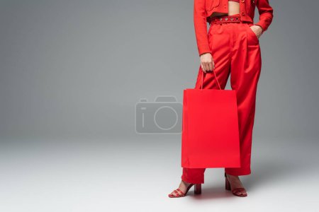 cropped view of woman holding shopping bag while standing with hand in pocket of red pants on grey background