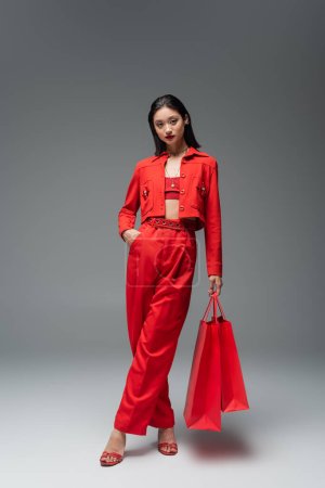 Foto de Longitud completa de la elegante mujer asiática posando con bolsas de compras y la mano en el bolsillo de los pantalones rojos sobre fondo gris - Imagen libre de derechos