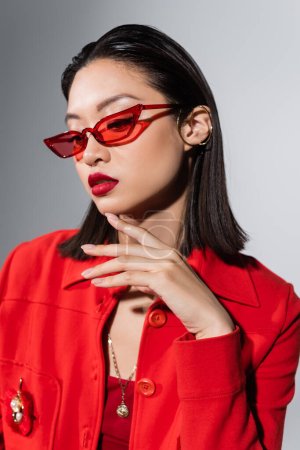 Porträt einer asiatischen Frau mit roter stylischer Sonnenbrille und Ohrenmanschette, die das Kinn isoliert auf grau berührt