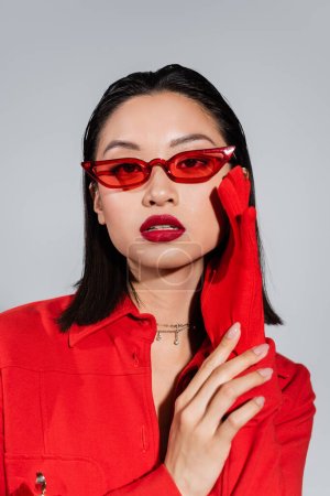 portrait of asian woman in fashionable sunglasses and red glove holding hand near face isolated on grey