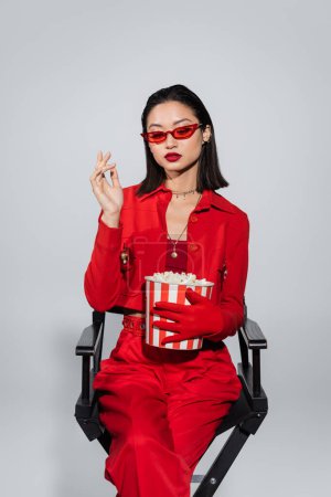 brünette asiatische Frau in stylischer Sonnenbrille und rotem Anzug sitzt mit Eimer Popcorn isoliert auf grau