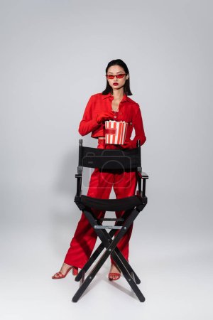 modische asiatische Frau mit roter Sonnenbrille hält Eimer Popcorn in der Nähe Stuhl auf grauem Hintergrund