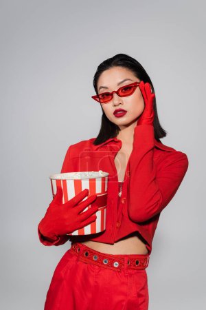 Téléchargez les photos : Asiatique femme en rouge gants tenant pop-corn et ajustement à la mode lunettes de soleil isolé sur gris - en image libre de droit