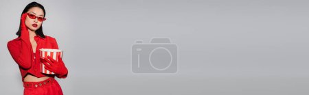 Téléchargez les photos : Femme asiatique en vêtements à la mode tenant seau de pop-corn et réglage des lunettes de soleil rouges isolées sur gris, bannière - en image libre de droit