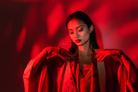 elegant asian woman in gloves and kimono cape posing on red background with shadow