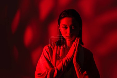 jeune femme asiatique en kimono cape debout avec des mains priantes sur fond sombre avec de la lumière rouge