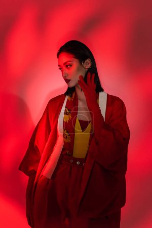 fashionable asian woman in kimono cape and glove touching face on abstract background with red light