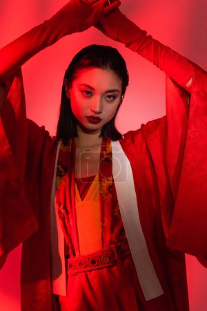 mujer asiática en kimono capa y guantes posando con las manos por encima de la cabeza sobre fondo rojo