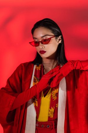 asiatisches Model in Kimono-Umhang und modischer Sonnenbrille posiert in Handschuhen auf rotem Hintergrund