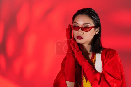 Foto de Mujer asiática de moda en guantes rojos y gafas de sol de moda cogidas de la mano cerca de la cara sobre fondo rojo abstracto - Imagen libre de derechos