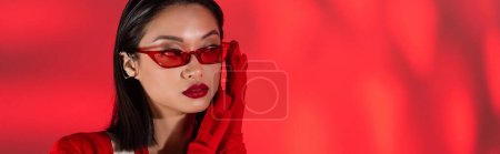 Porträt einer asiatischen Frau mit trendiger Sonnenbrille und Handschuhen auf rotem abstrakten Hintergrund, Banner