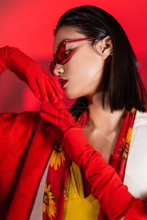 sensual mujer asiática en gafas de sol y pañuelo con estampado floral posando en guantes sobre fondo rojo