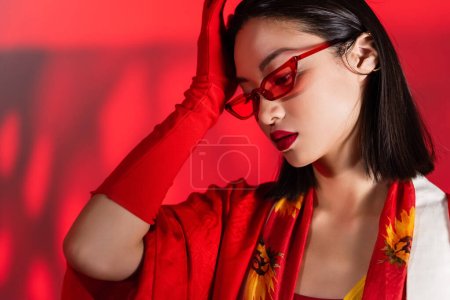 trendige asiatische Frau in Sonnenbrille und Schal mit floralem Print, der die Stirn auf rotem Hintergrund berührt