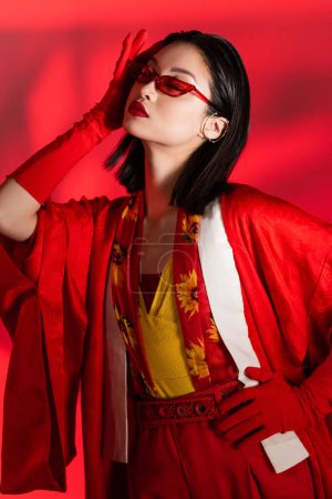 femme asiatique en kimono cape et lunettes de soleil posant avec la main sur la hanche et les yeux fermés sur fond rouge avec ombre