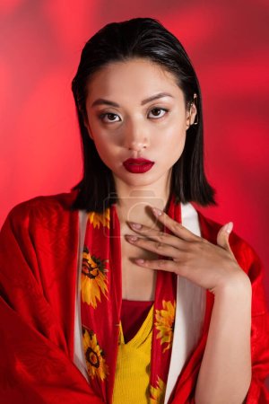 brünette asiatische Frau in Kimono-Umhang und Schal mit floralem Dekor schaut in die Kamera auf abstraktem rotem Hintergrund
