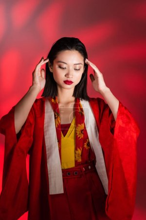 elegante asiatische Frau in rotem Kimono-Umhang posiert mit den Händen in der Nähe des Kopfes auf abstraktem Hintergrund