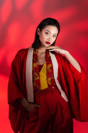 elegante asiatische Frau in Kimono-Umhang und Schal mit floralem Print stehend mit der Hand in der Tasche auf rotem Hintergrund