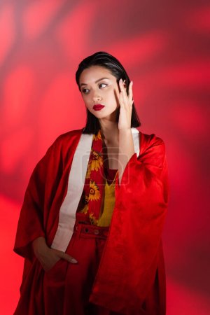 elegante asiatische Frau im Kimono-Umhang, die das Gesicht berührt, während sie mit der Hand in der Tasche auf rotem abstrakten Hintergrund steht