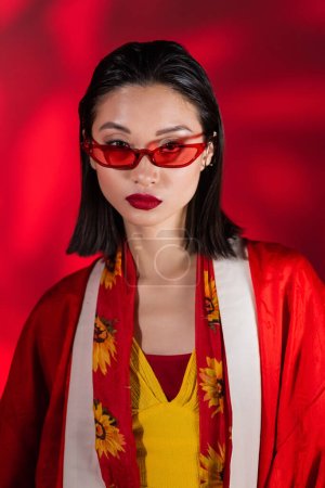 Porträt einer asiatischen Frau mit stylischer Sonnenbrille und Kimono-Umhang, die auf abstraktem rotem Hintergrund in die Kamera blickt