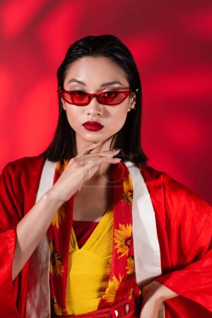 asiatische Frau in trendiger Sonnenbrille und Kimono-Umhang mit floralem Print-Schal, der das Kinn auf abstrakt rotem Hintergrund berührt 