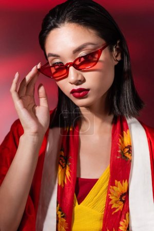 Photo for Portrait of brunette asian woman touching red sunglasses and looking at camera on dark background - Royalty Free Image