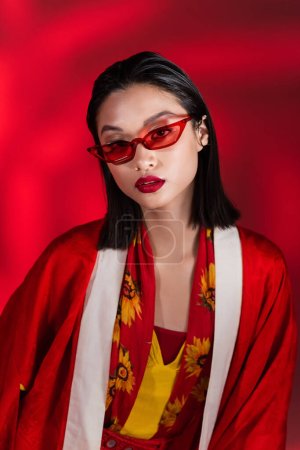 Photo for Brunette asian woman in elegant kimono cape and stylish sunglasses looking at camera on red background - Royalty Free Image