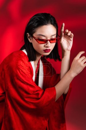 sinnliche asiatische Frau in Kimono-Umhang und trendiger Sonnenbrille posiert auf rotem Hintergrund