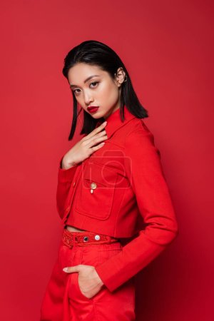 young asian woman in stylish outfit touching chest while holding hand in pocket on red background