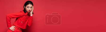 Photo for Stylish asian woman in elegant attire posing with hand on hip while touching face on red background, banner - Royalty Free Image
