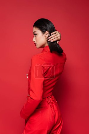 Foto de Vista lateral de morena mujer asiática en traje elegante fijación de pelo mientras mira hacia otro lado sobre fondo rojo - Imagen libre de derechos