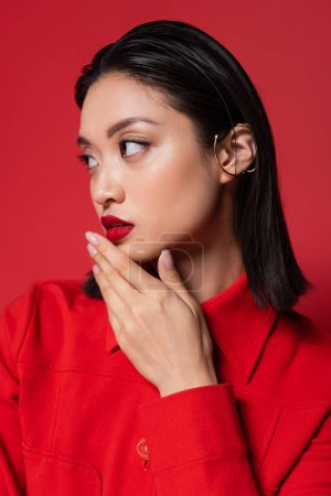 Foto de Retrato de morena mujer asiática con el manguito de la oreja y el maquillaje de la mano cerca de la barbilla mientras mira hacia otro lado aislado en rojo - Imagen libre de derechos